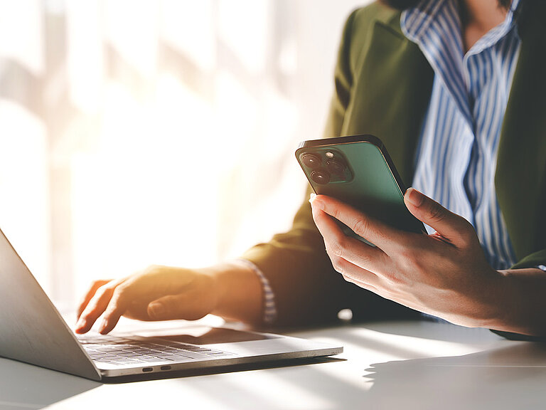 Person sitzt an einem Schreibtisch und nutzt gleichzeitig ein Smartphone und einen Laptop, mit Sonnenlicht auf den Händen und der Tastatur