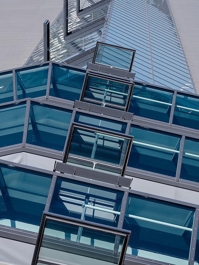 Schräg verlaufendes Glasdach PR60 mit mehreren Lüftungselementen auf moderner Gebäudestruktur.