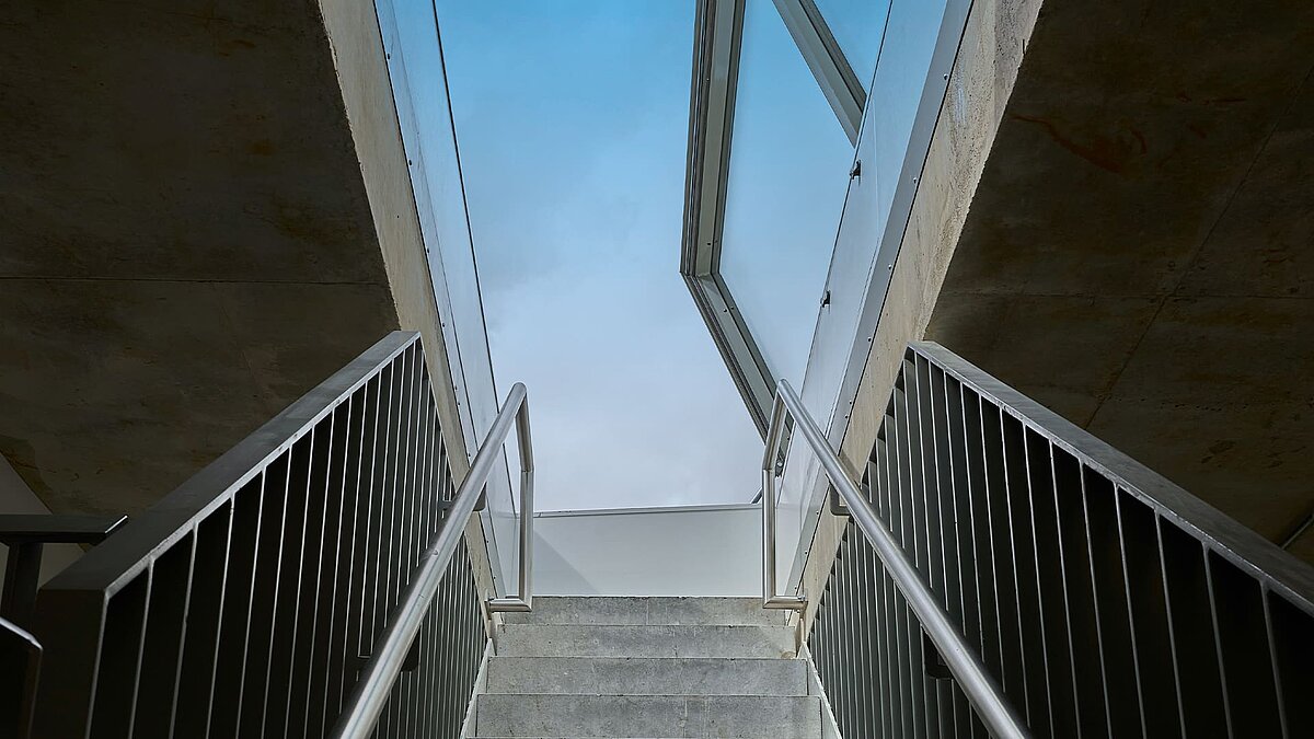 Blick von unten auf eine Betontreppe mit Metallgeländern, die zu einem geöffneten LAMILUX Flachdach Ausstieg führt.