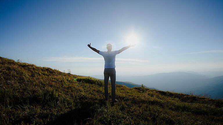 erson steht mit ausgebreiteten Armen auf einem Hügel in der Natur, Blick in den Himmel bei Sonnenlicht