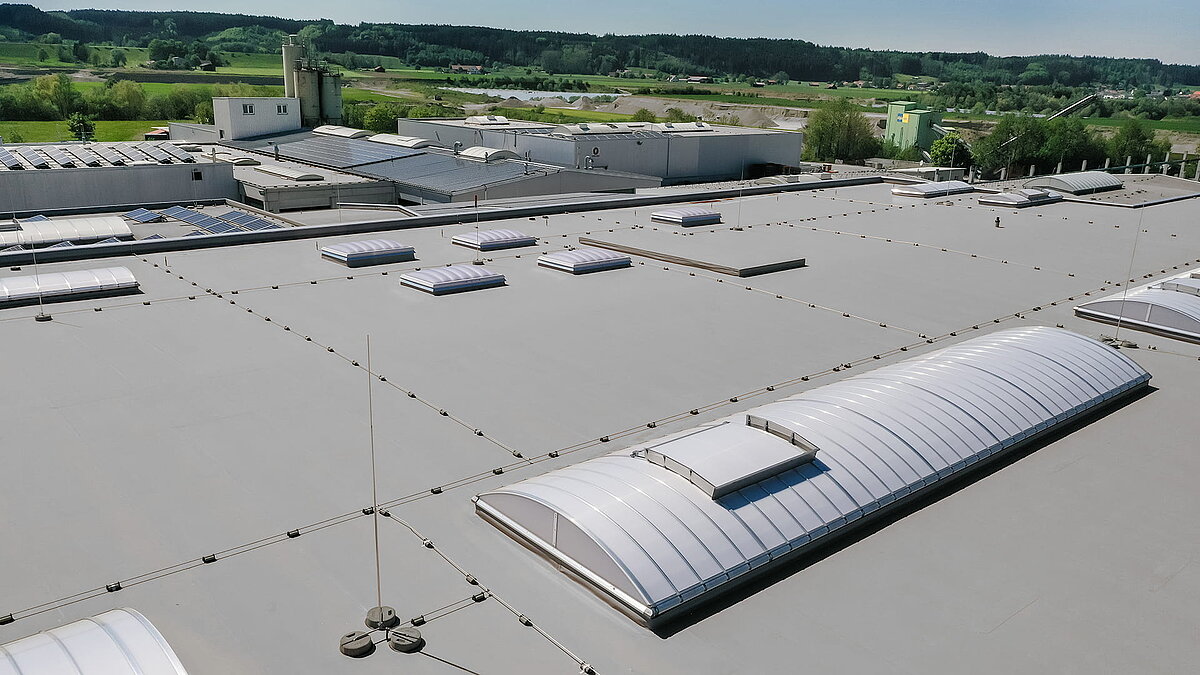 Dach der Produktionshalle des Bauunternehmens Unglehrt mit LAMILUX Lichtkuppeln F100 W und Lichtbändern B, umgeben von grüner Landschaft.