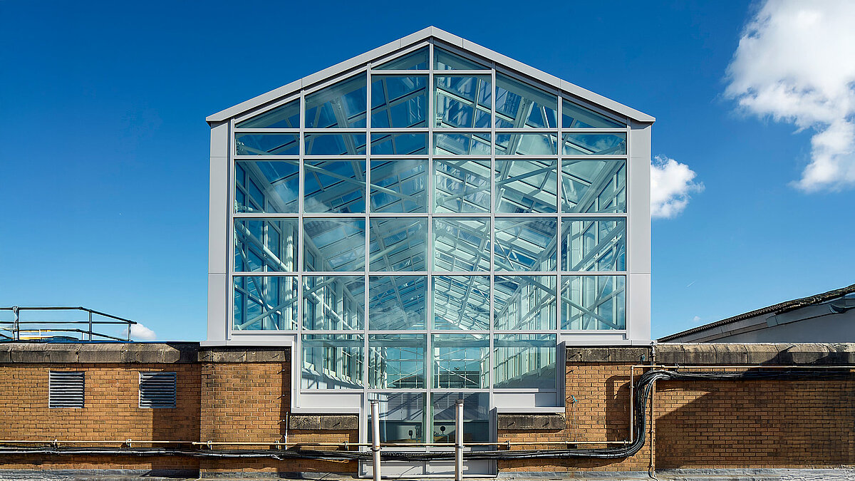 Modernes Glasdach mit Stahlrahmen auf einem Industriegebäude, blauer Himmel und Wolken reflektieren im Glas