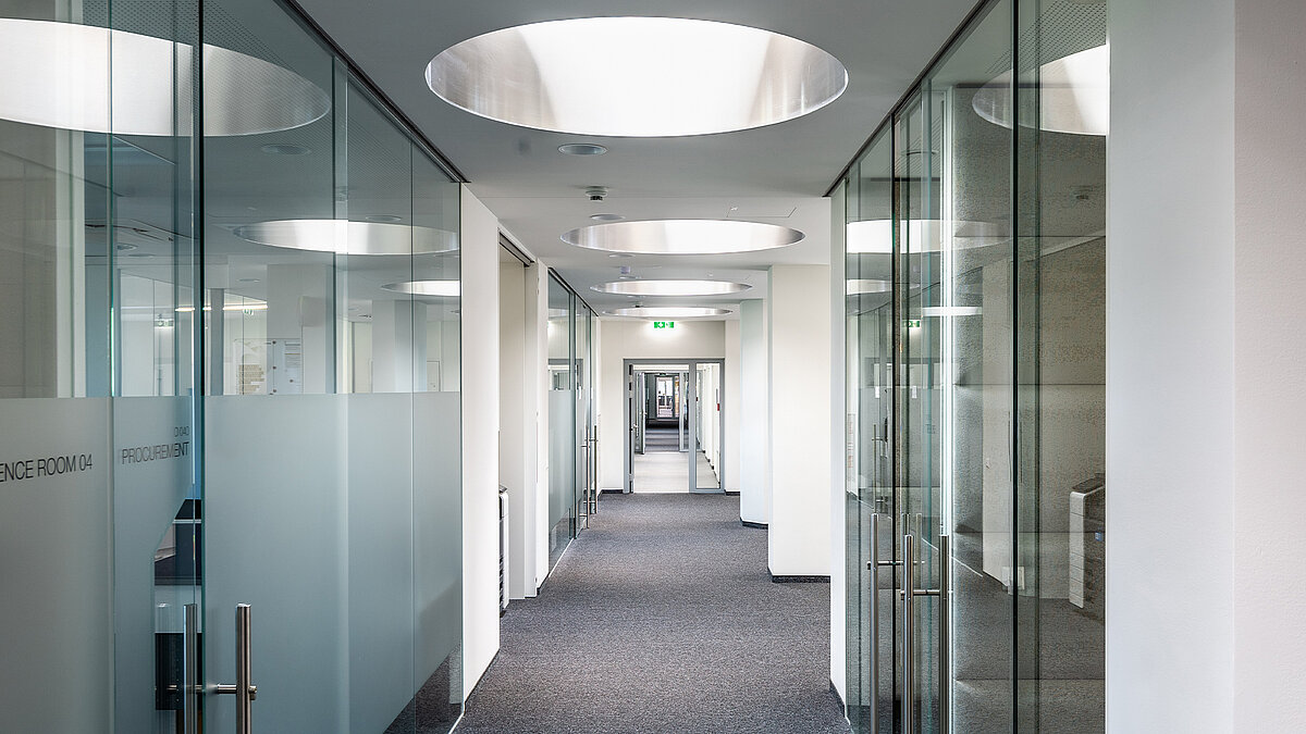 Blick in einen Bürogang mit Glaswänden links und rechts und runden Flachdach Fenstern für Tageslichteinfall.