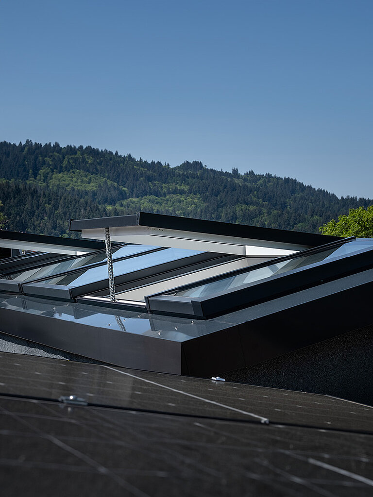 Modulares Glasdach MS78 mit geöffneten Lüftungselementen auf Flachdach vor Waldkulisse.