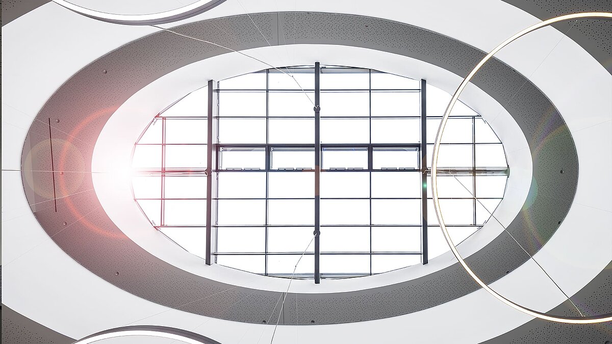 View upwards to an oval glass roof with sunlight and modern ring lamps in the atrium of an office building.