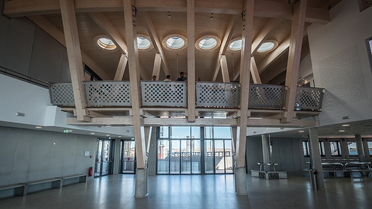 Innenraum des Hafenterminals Norderney mit sichtbaren Holzbalken, runden Flachdach Fenstern und modernem Geländer.