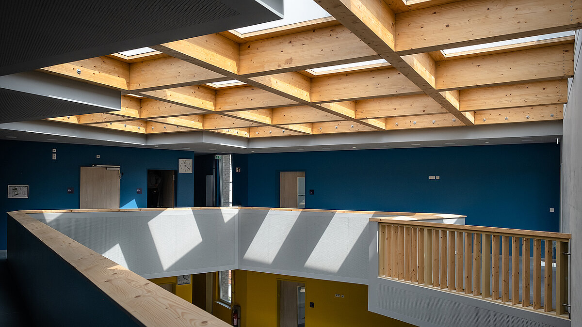 Atrium eines modernen Schulgebäudes mit Holzbalkendecke, Flachdach Fenstern, blauer Wand und Holzgeländer