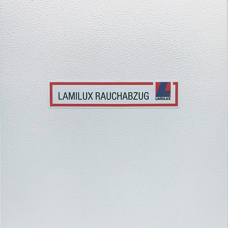 Beschriftetes Hinweisschild mit der Aufschrift „LAMILUX RAUCHABZUG“ auf strukturierter Wandoberfläche