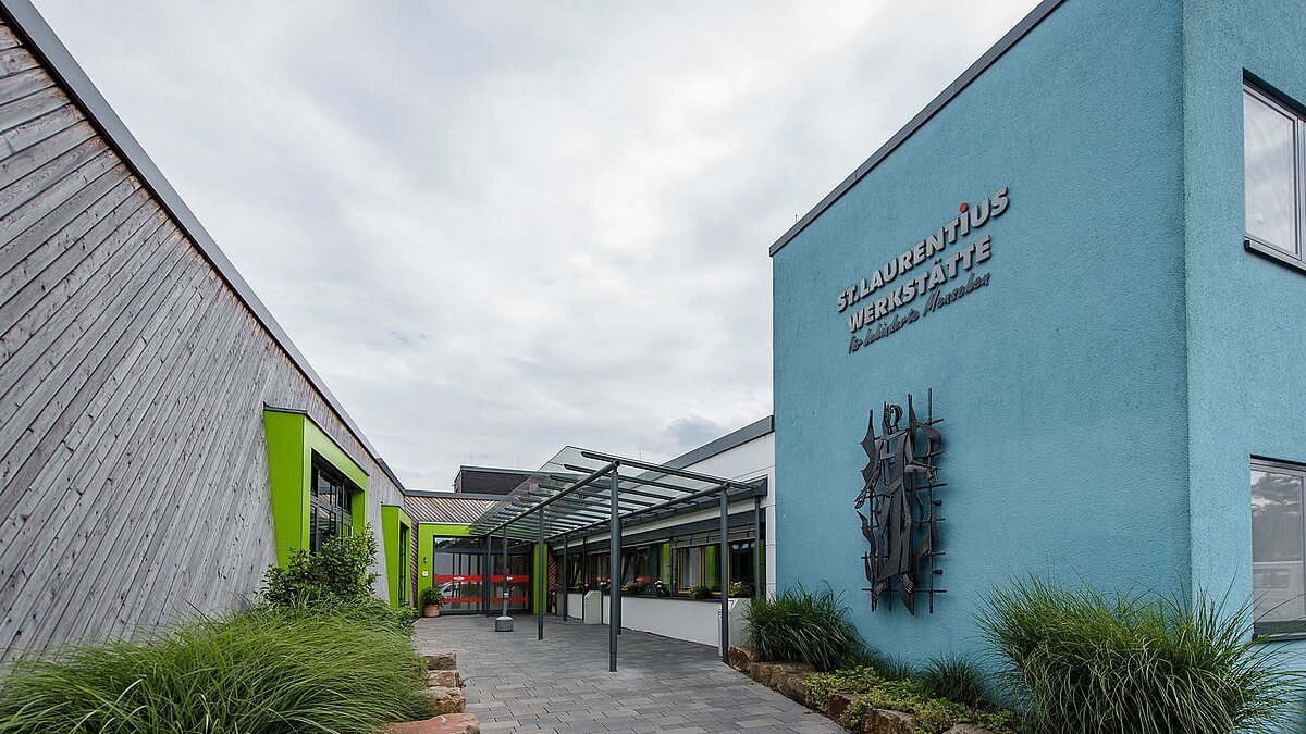 Eingangsbereich der St. Laurentius Werkstatt des Caritasverbandes in Hagen mit Glasvordach, blauer Fassade und grünen Fensterrahmen, umgeben von Pflanzen.