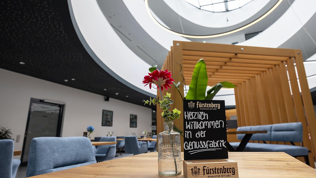 Innenaufnahme eines modernen Gastraums mit runden Oberlichtern, Holzelementen und einer Tafel mit der Aufschrift „Herzlich willkommen in der Genussfabrik“.