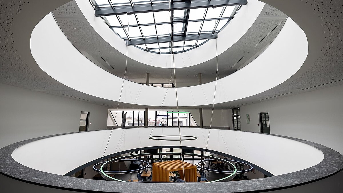 Modernes Atrium mit ovalem Lichthof und Glasdach, runde LED-Leuchten und offener Blick auf den Arbeitsbereich im Erdgeschoss.