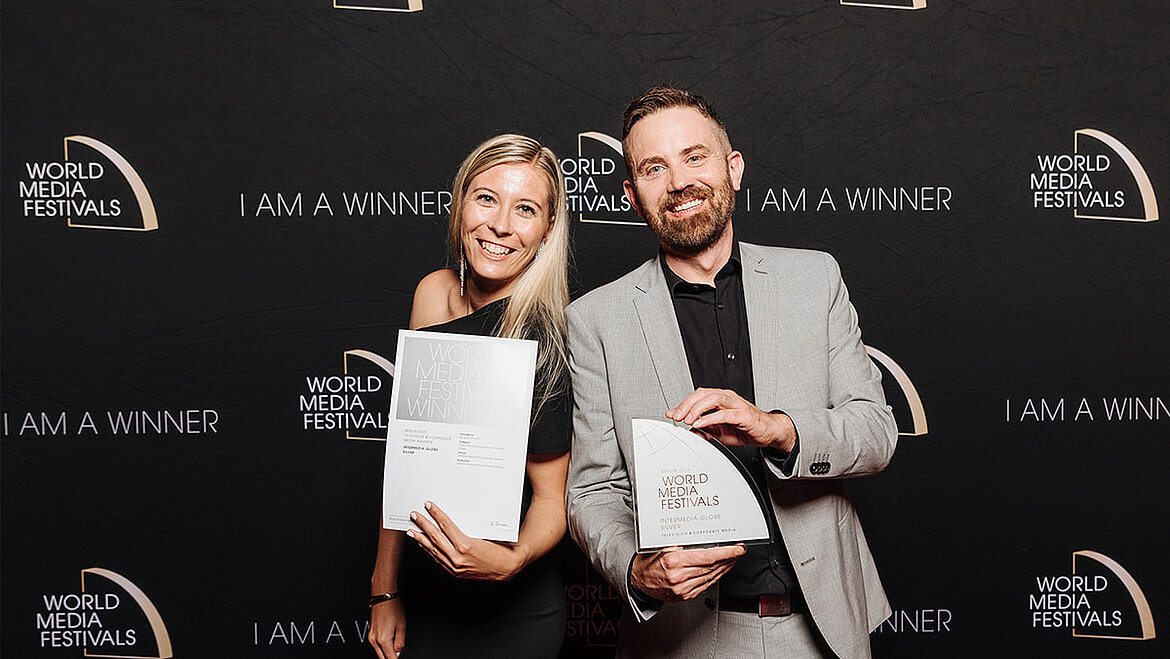 Zwei Personen stehen nebeneinander vor einer dunklen Veranstaltungswand und halten eine Auszeichnung sowie eine gerahmte Urkunde in den Händen