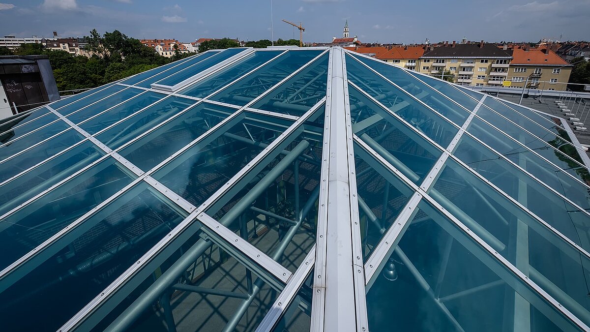 Detailaufnahme des Glasdachs des Dante-Gymnasiums mit blauer Verglasung und stabiler Stahlkonstruktion, darunter sichtbare Rohre und Träger.
