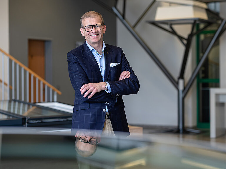 Person mit Brille und verschränkten Armen in modernem Gebäudeinnenraum mit Stahlkonstruktion und Galerie im Hintergrund