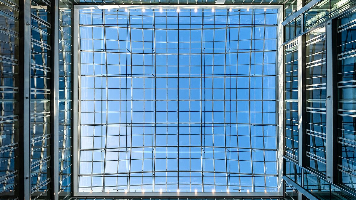 Blick nach oben auf das Glasdach des Atriums im Bürogebäude Newton der TÜV Süd Gruppe in München, mit blauem Himmel und modernen Glasstrukturen.