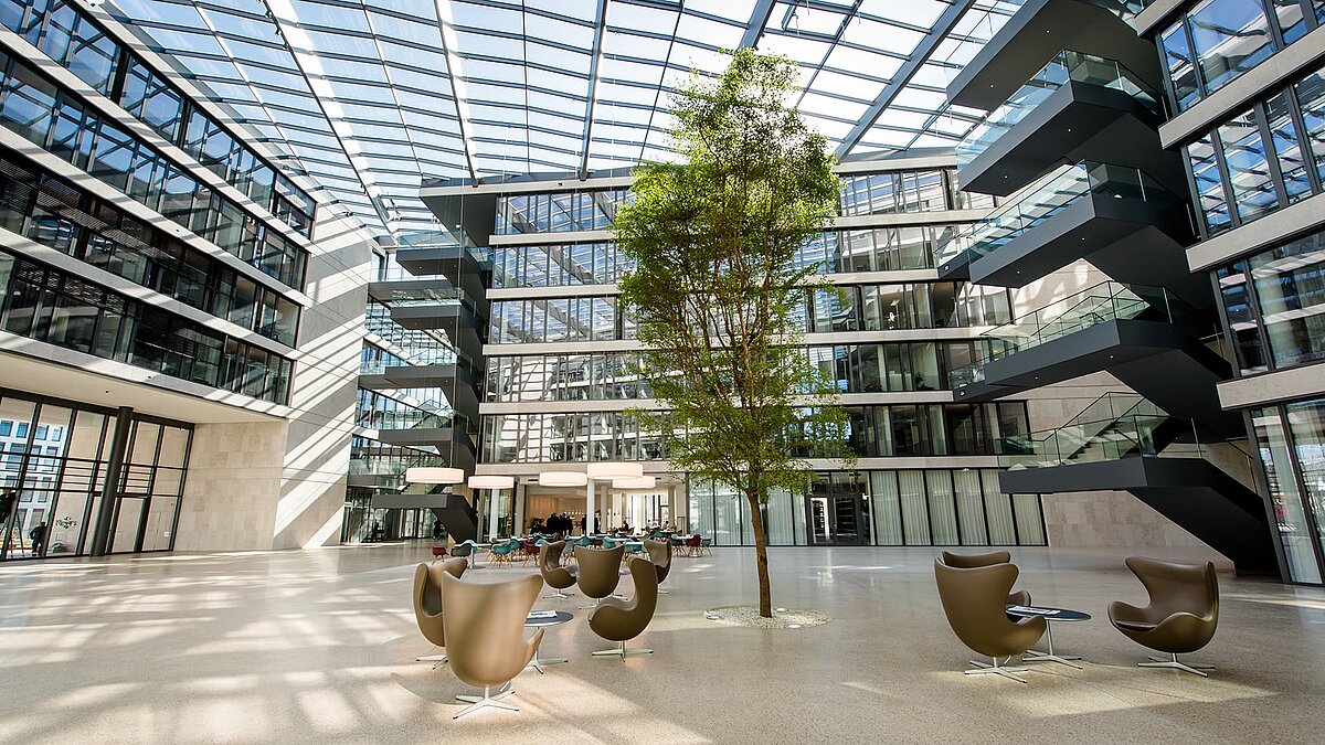 Atrium des FGS Campus mit Glasdach, moderner Innenarchitektur, einer zentralen grünen Baumfläche und eleganter Sitzgelegenheit.