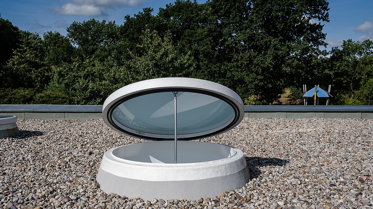 Geöffnetes rundes Oberlicht auf dem Dach der Kindertagesstätte Sonnenfeld in Wandlitz, umgeben von Kies und mit grünem Baumbestand im Hintergrund.