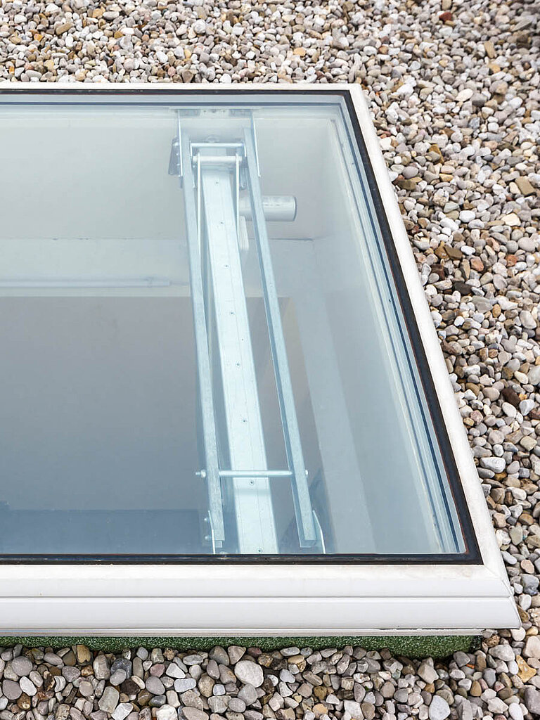 Detailansicht eines LAMILUX Flachdach Fenster F100 RWA auf Kies-Flachdach mit transparenter Verglasung und sichtbarer Antriebstechnik