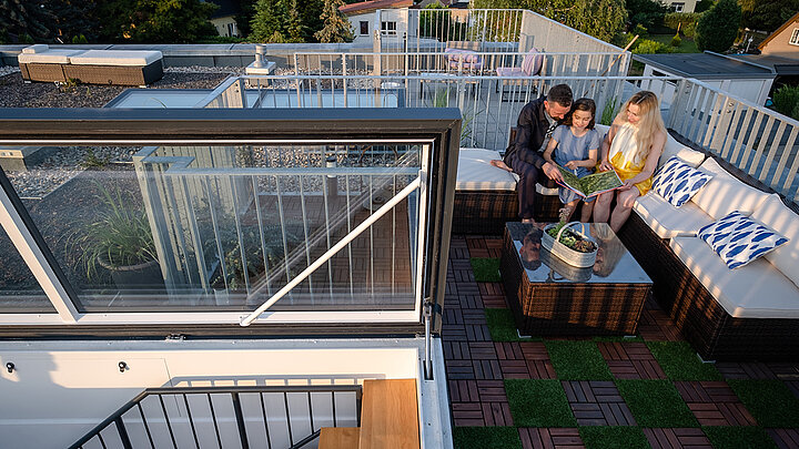 Familie sitzt auf einer modernen Dachterrasse mit Lounge-Möbeln, umgeben von Pflanzen, mit Zugang über einen Flachdachausstieg.