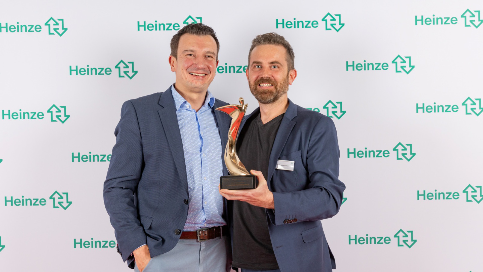 Zwei Männer in Anzügen stehen vor einer Veranstaltungswand und halten gemeinsam eine Auszeichnung in den Händen